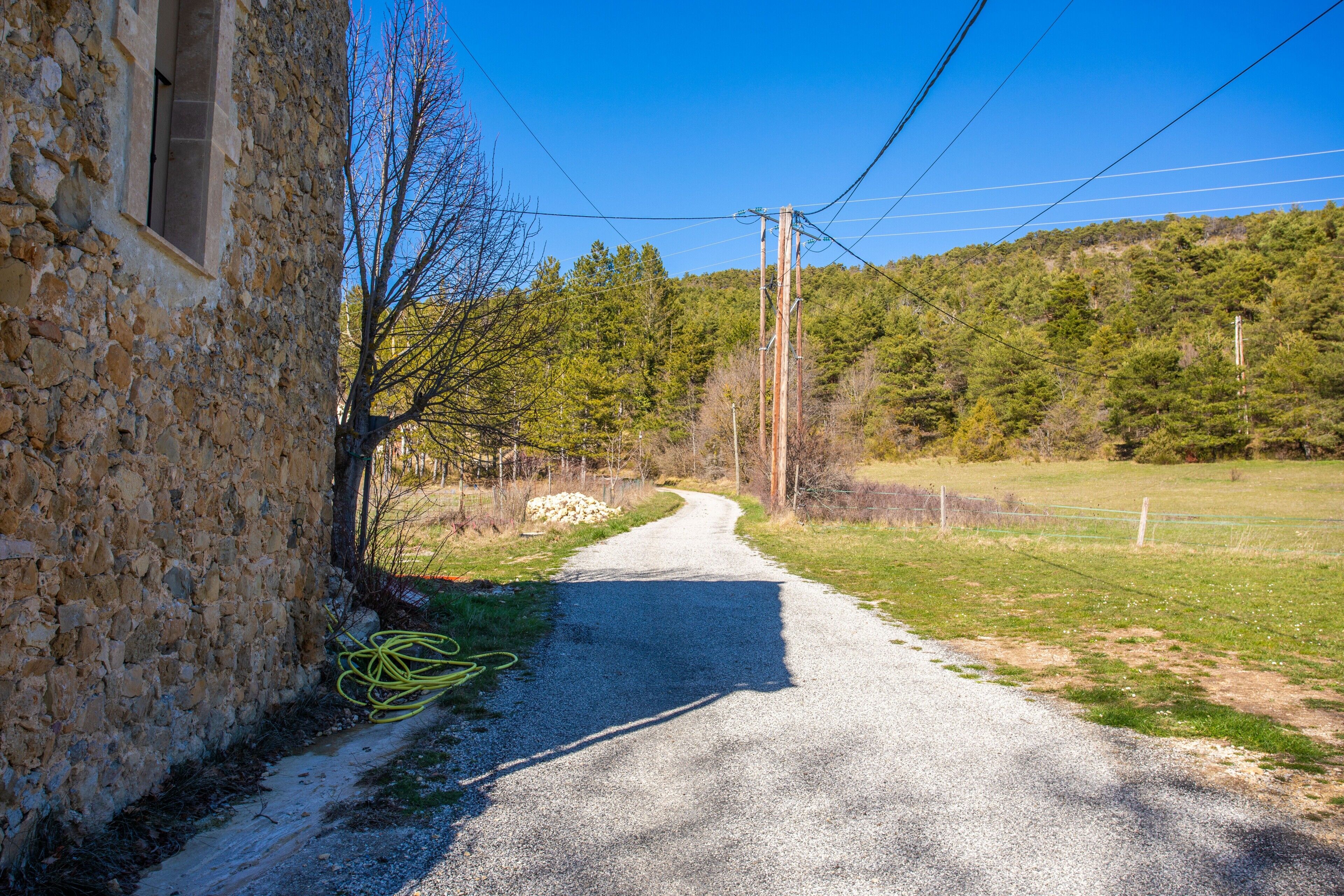 Una strada rurale con un edificio in pietra a sinistra, immerso tra alberi e un prato.