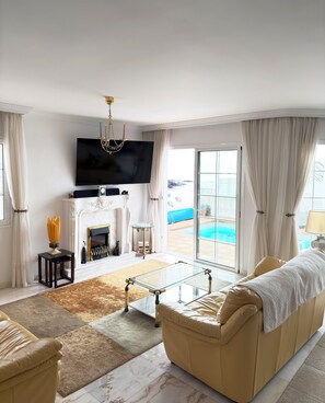 Ein Wohnzimmer mit Sofa, Glastisch, Kamin, Fernseher und Blick auf den Pool.