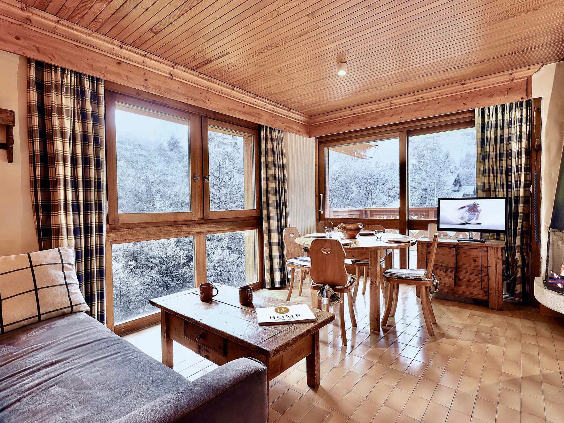 A cozy room with a wooden ceiling, large windows with plaid curtains, a wooden dining table, and a television.