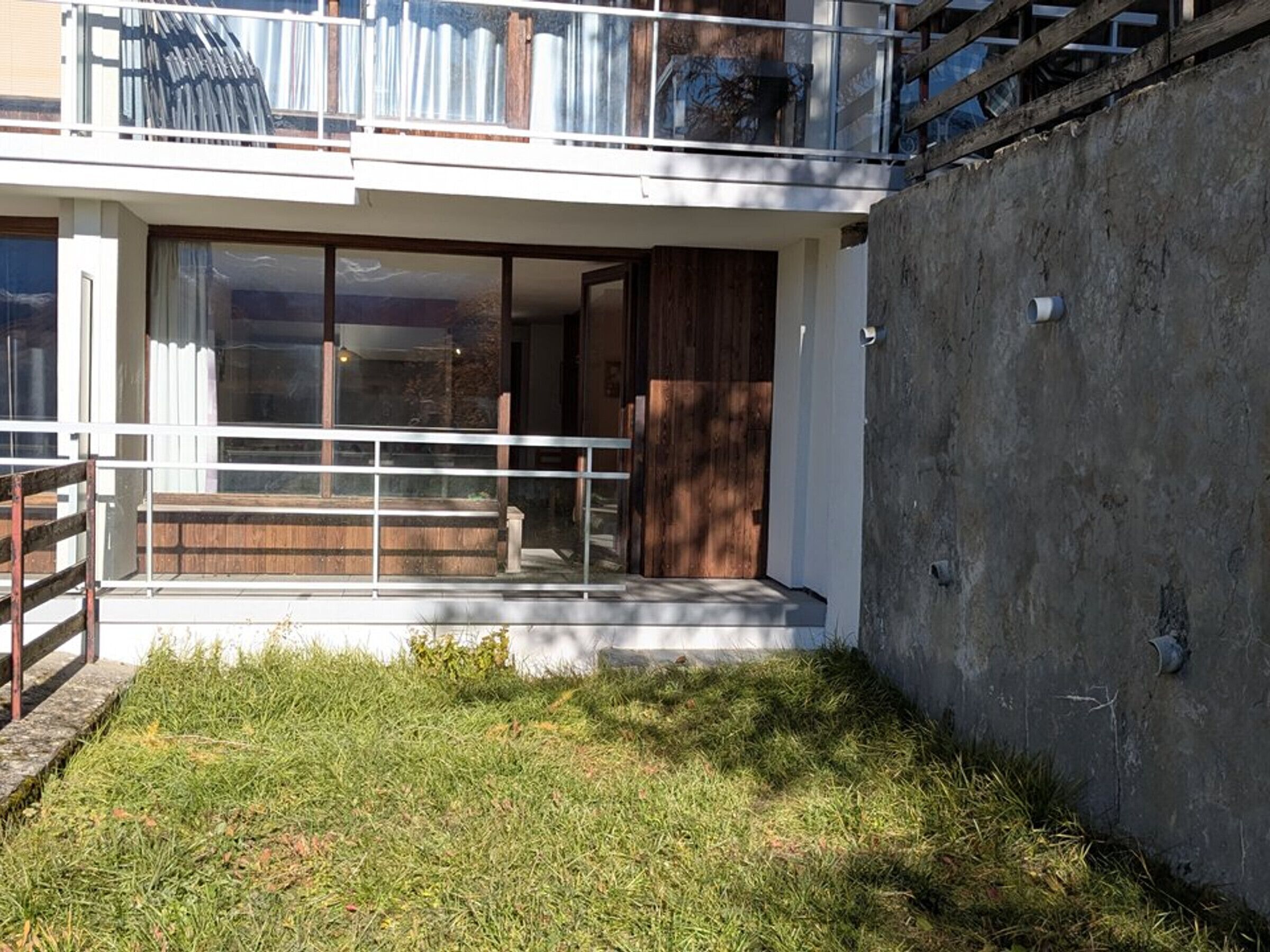 A modern two-story house with a glass door entrance, a wooden deck, and a concrete wall.