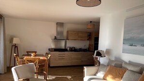 Un soggiorno moderno con divano, tavolo da pranzo con sedie, angolo cottura con mobili in legno e un'ampia finestra vista mare.