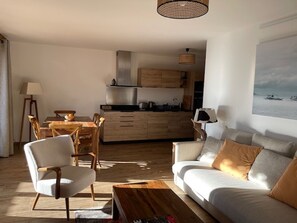 Ein modernes Wohnzimmer mit einer Couch, einem Esstisch mit Stühlen, einer Küchenzeile mit Holzschränken und einem großen Fenster mit Meerblick.