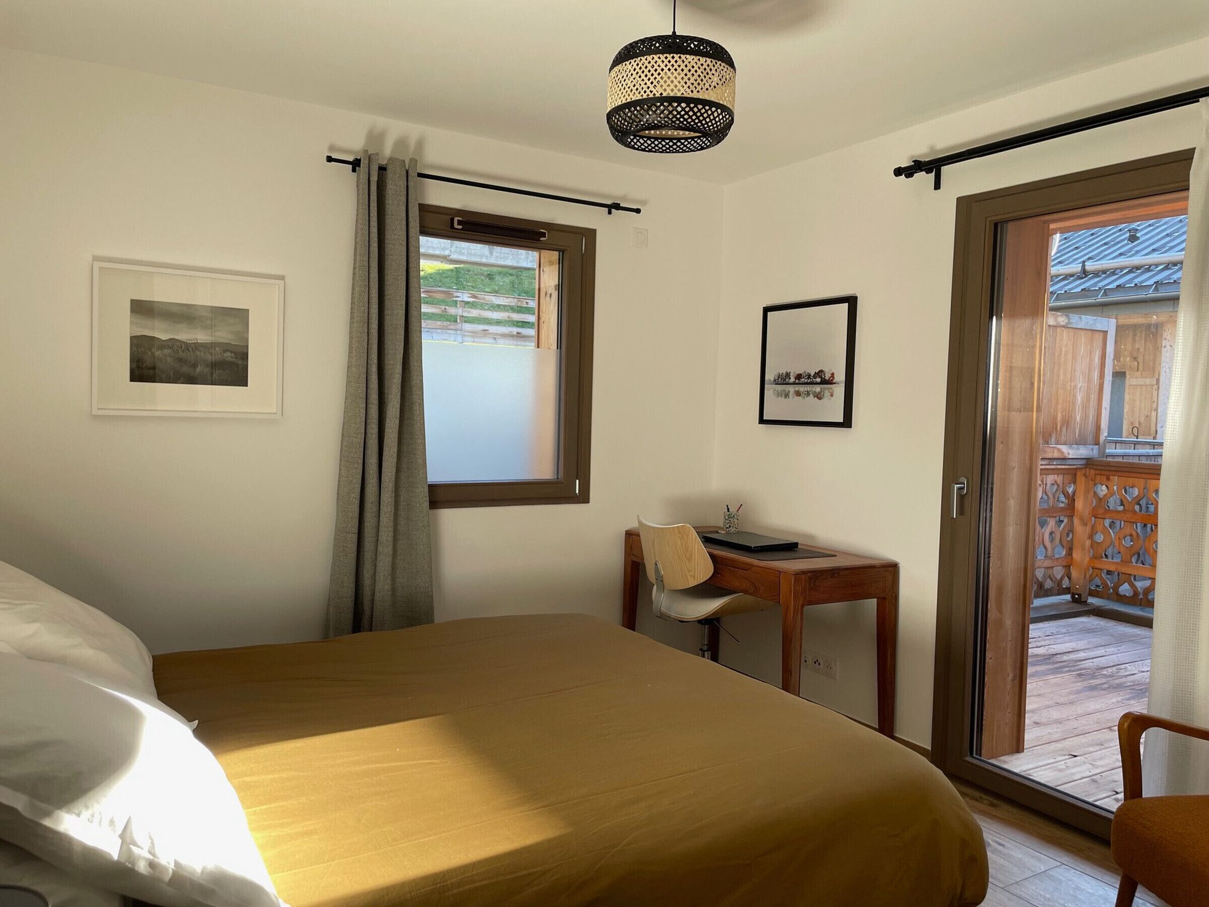 Une chambre à coucher avec un lit, un bureau et une chaise. Il y a une fenêtre avec des rideaux, un tableau au mur et une porte qui donne sur un espace extérieur.