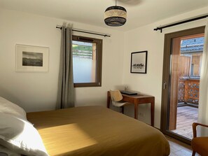 Ein Schlafzimmer mit Bett, Schreibtisch und Stuhl. Ein Fenster mit Vorhängen, ein Bild an der Wand und eine Tür zu einem Außenbereich.