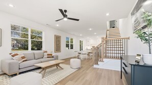 Uma sala de estar moderna com sofá, mesa de centro e um banquinho, uma escadaria que leva a uma cozinha e janelas amplas com persianas.