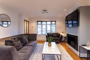 Ein modernes Wohnzimmer mit einem grauen Sofa, einem weißen Couchtisch mit Pflanze, einem Kamin und einem großen, an der Wand befestigten Fernseher.