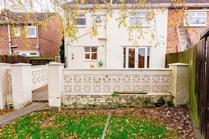 A residential property with a brick exterior, a white fence, and a small garden area with grass and fallen leaves. - Toast Stays - Gilmonby Grove (Middlesbrough)