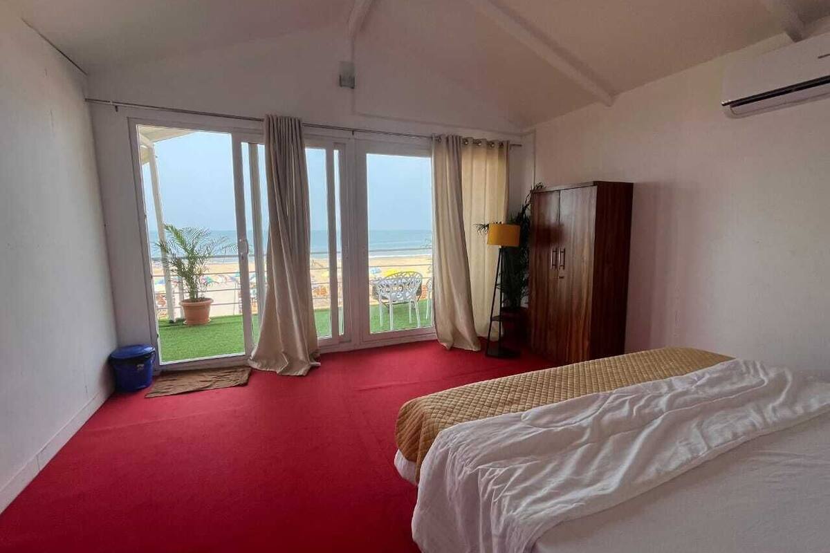 A hotel room with a red carpet, a bed, a wardrobe, and a view of the beach through large windows.