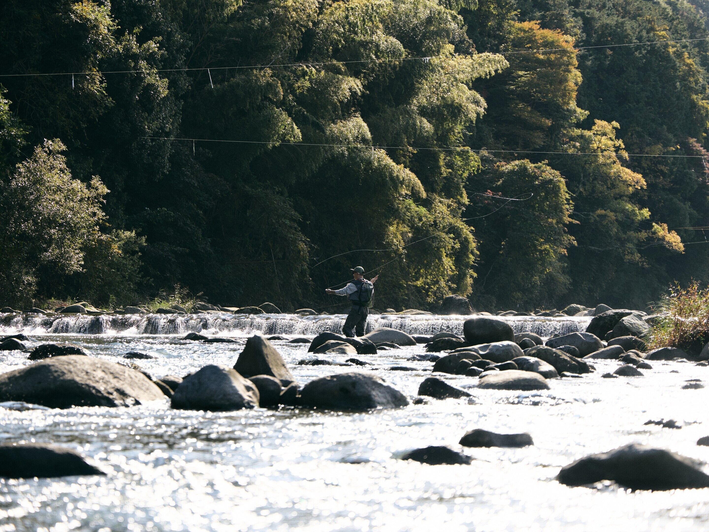 Una persona pescando en un río con rocas y árboles al fondo.