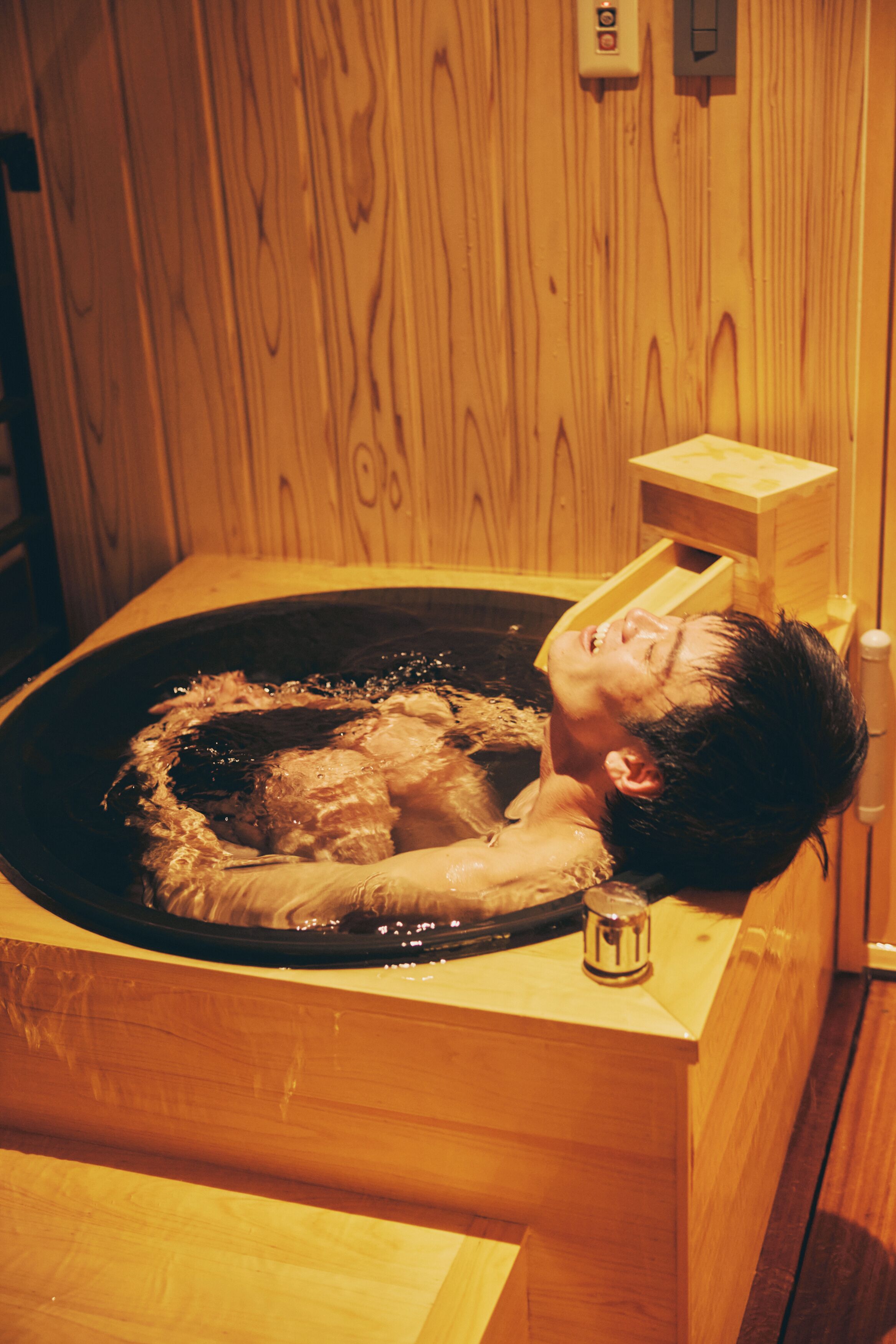 A wooden bathtub with a person lying inside, a wooden wall, and a small wooden cabinet.