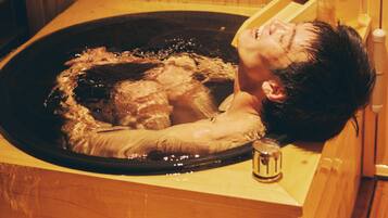 A wooden bathtub with a person lying inside, a wooden wall, and a small wooden cabinet.
