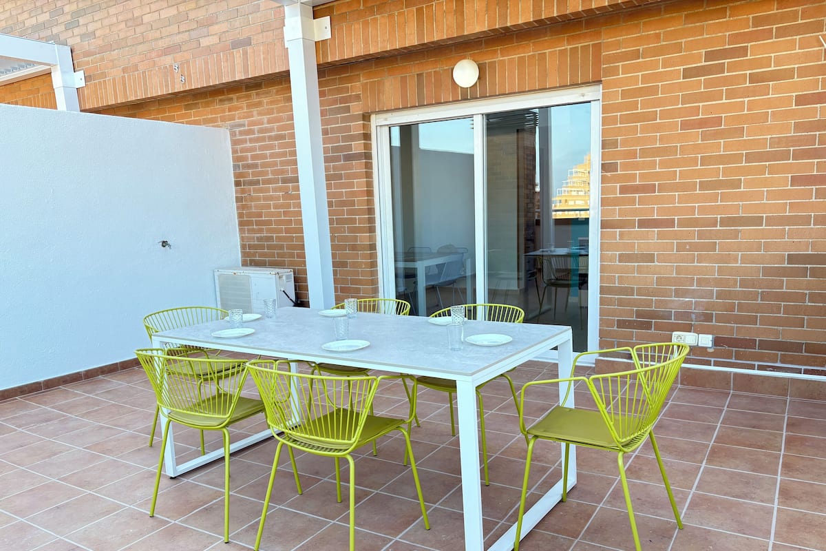 Een terras buiten met een witte tafel en gele stoelen, een bakstenen muur en een schuifdeur van glas.