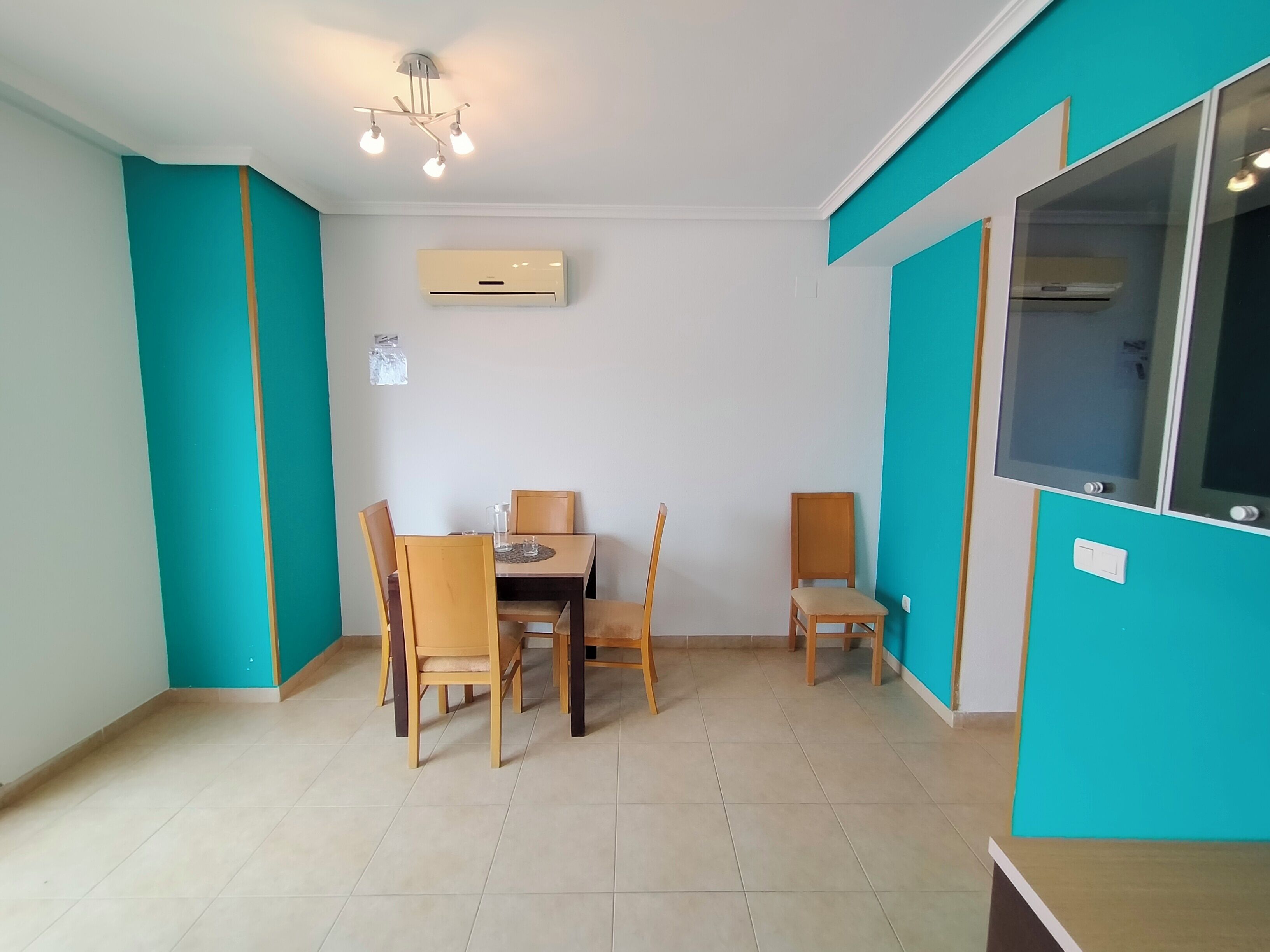 A room with teal walls, a dining table with chairs, a ceiling light fixture, and an air conditioning unit.