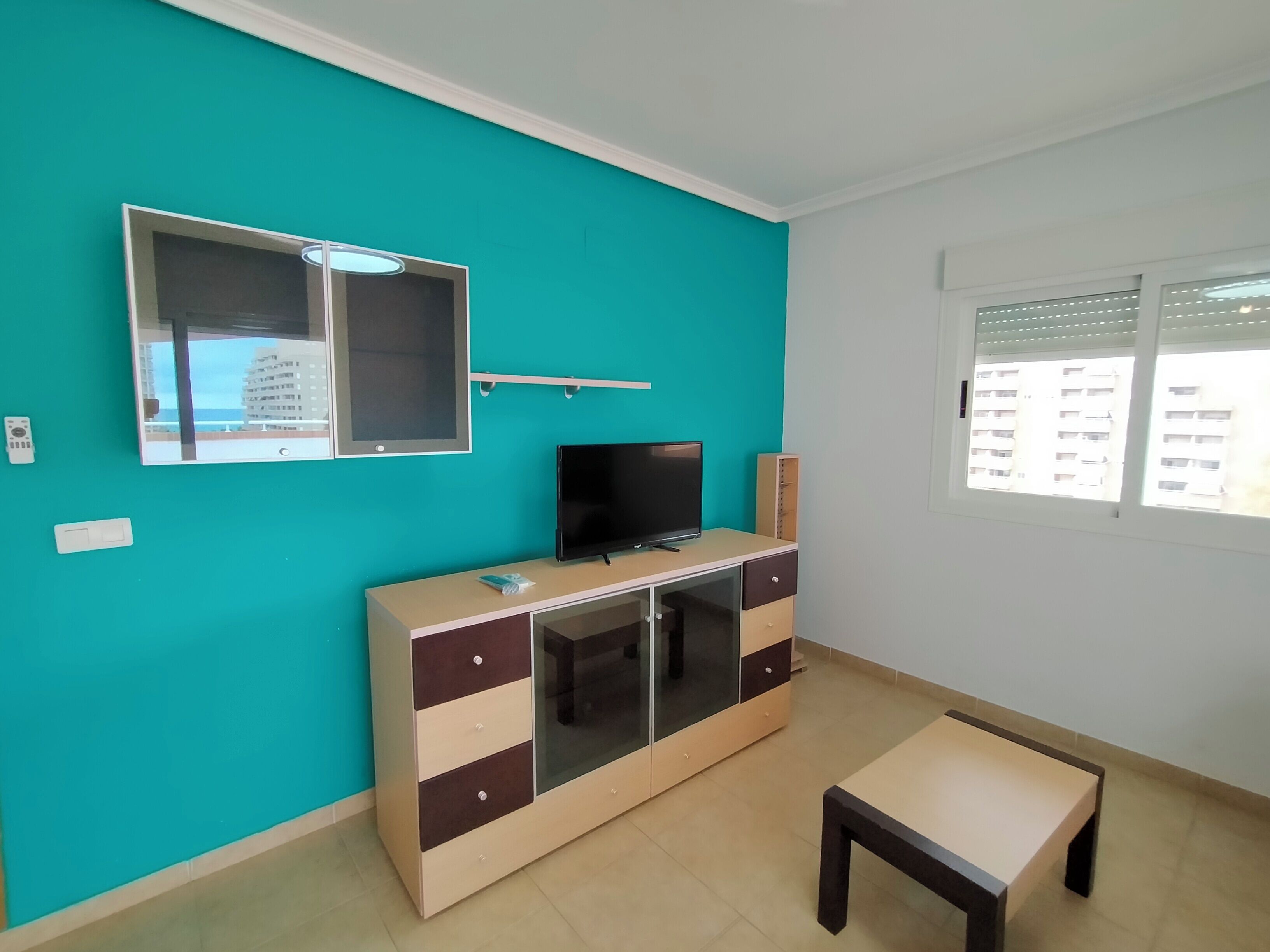 Una habitación con paredes color turquesa, un televisor sobre un mueble, una ventana con vistas a edificios y una mesa pequeña.