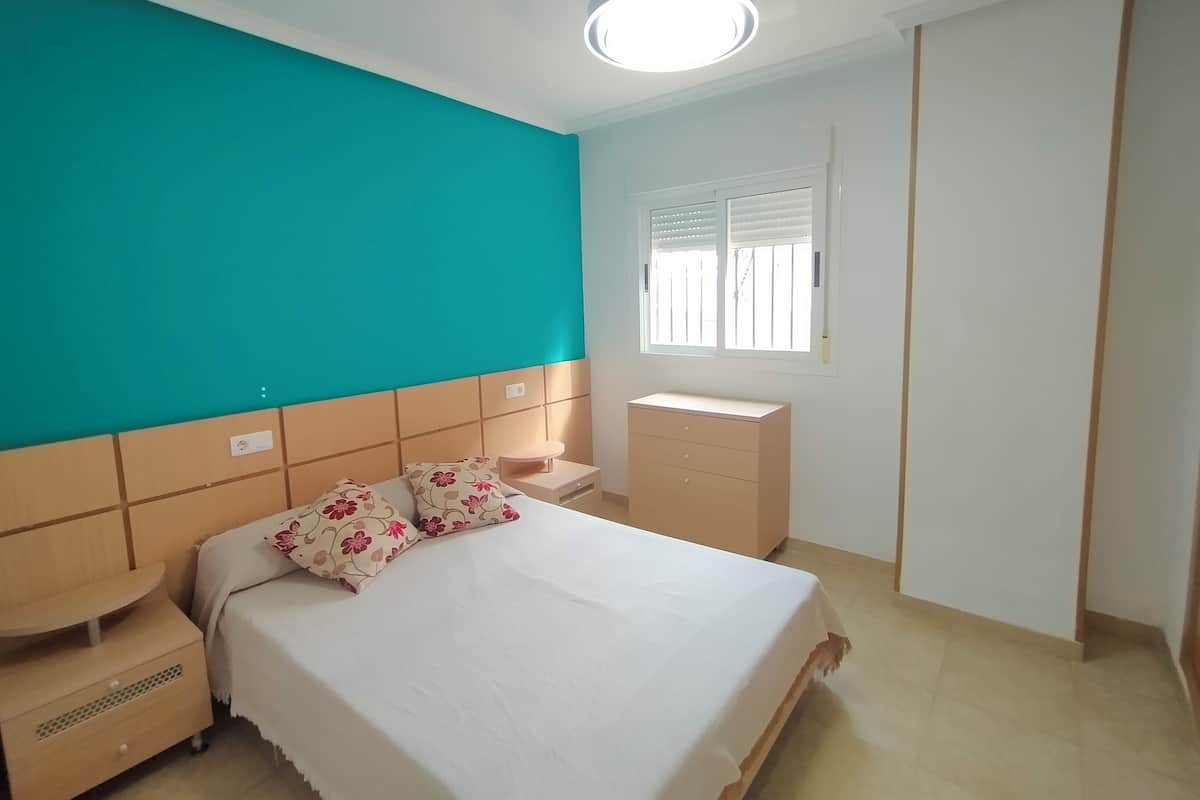 Een slaapkamer met een bed, nachtkastjes, een dressoir, een raam met jaloezieën en een wand in een blauwgroene tint.