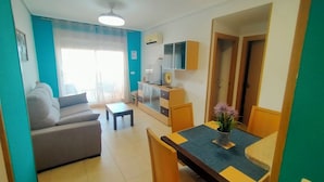 A modern living room with a sofa, a coffee table, and a dining area with a table and chairs. There is a television and a cabinet in the room. - ACV - Apartamentos Torremar (Oropesa del Mar)
