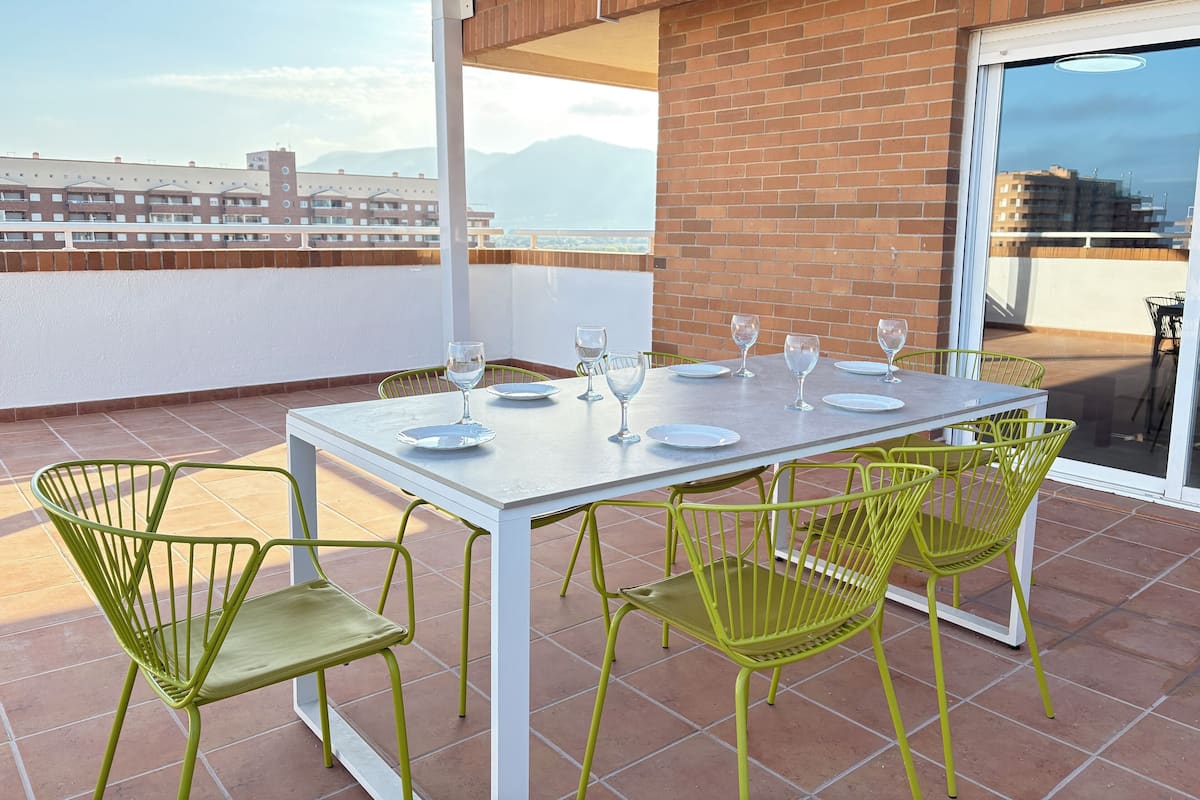 Een terras met een eettafel, groene stoelen en uitzicht op gebouwen en bergen.
