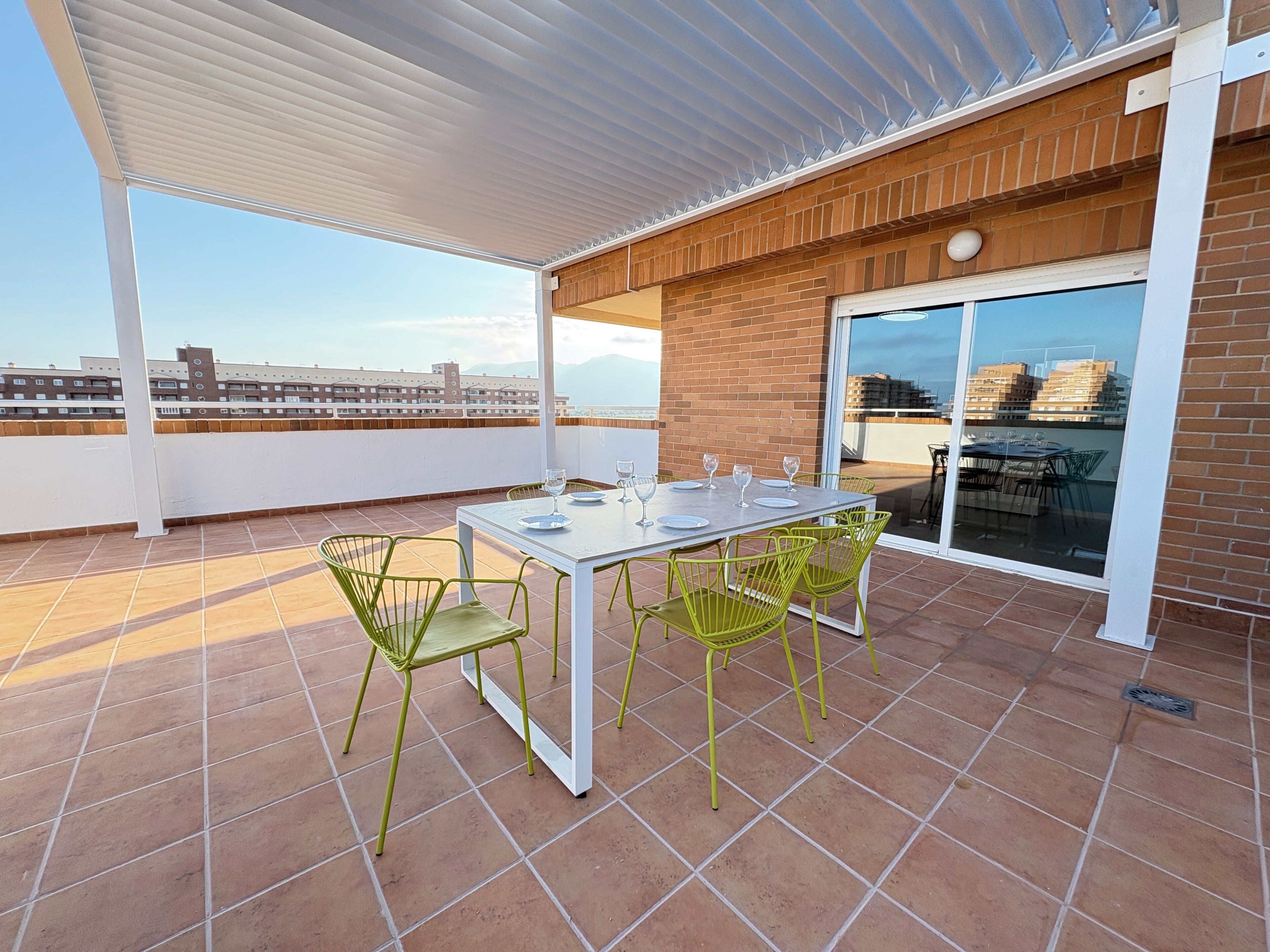 Eine überdachte Terrasse mit Tisch und Stühlen, im Hintergrund ein Blick auf Gebäude und eine Glastür zu einem weiteren Bereich.