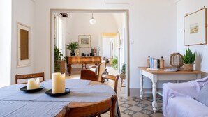 Un comedor con una mesa redonda, sillas y velas. Hay un sofá y una mesita con libros y una planta.