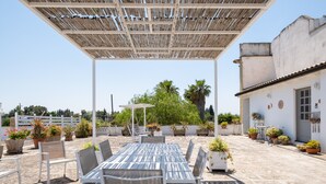Un patio exterior con mesa y sillas de madera, rodeado de plantas en macetas y una pérgola.