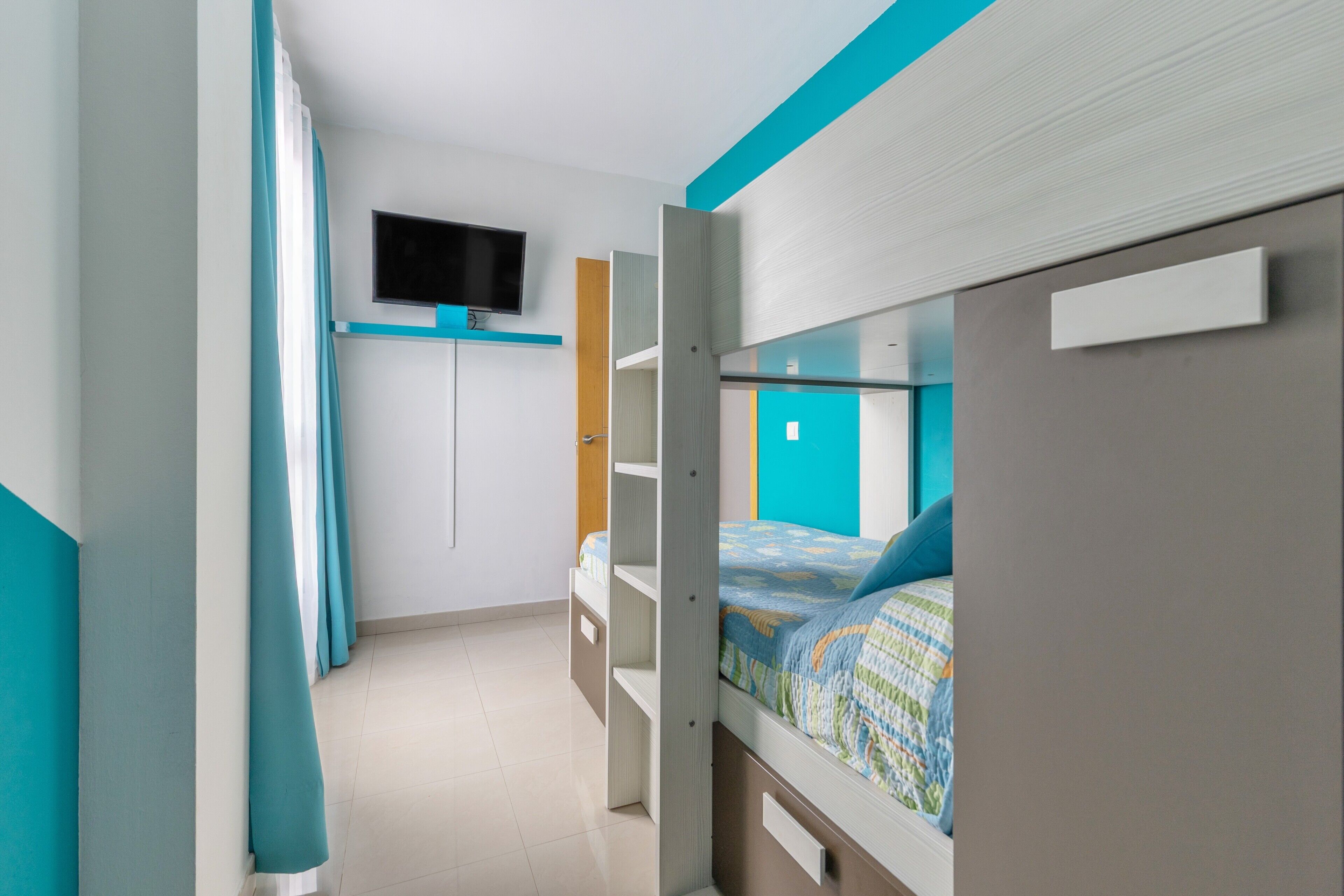 A bedroom with bunk beds, a TV mounted on the wall, and a window with curtains.