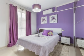 Un dormitorio con cama, mesitas de noche, espejo y ventana con cortinas moradas.