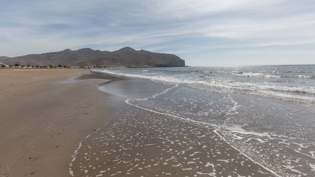 Ein Strand mit einem Berg im Hintergrund, einem klaren Himmel und ruhiger See.