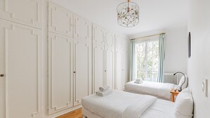 A bedroom with two beds, white walls, wooden flooring, a radiator, and a window with curtains. - Parc Montsouris - Dans un joli square au calme - 2 chambres (Paris)