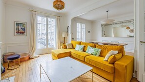 A bright living room with a yellow sofa, a white coffee table, and a large window with curtains. - Parc Montsouris - Dans un joli square au calme - 2 chambres (Paris)