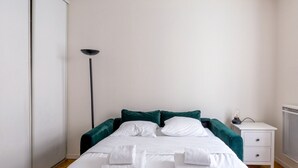 A neatly made bed with white linens and green pillows, a bedside table with a lamp, and a radiator on the wall. - Parc Monceau - Villiers - Studio (Paris)