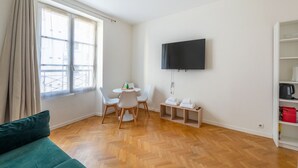 A modern living room with a wooden herringbone floor, a green sofa, a dining table with chairs, a flat-screen TV mounted on the wall, and a built-in bookshelf with a kettle and a red bag. - Parc Monceau - Villiers - Studio (Paris)