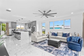 Una sala de estar moderna con sofá, reposapiés y sillón azul, una zona de cocina y un amplio ventanal con vista a una piscina.