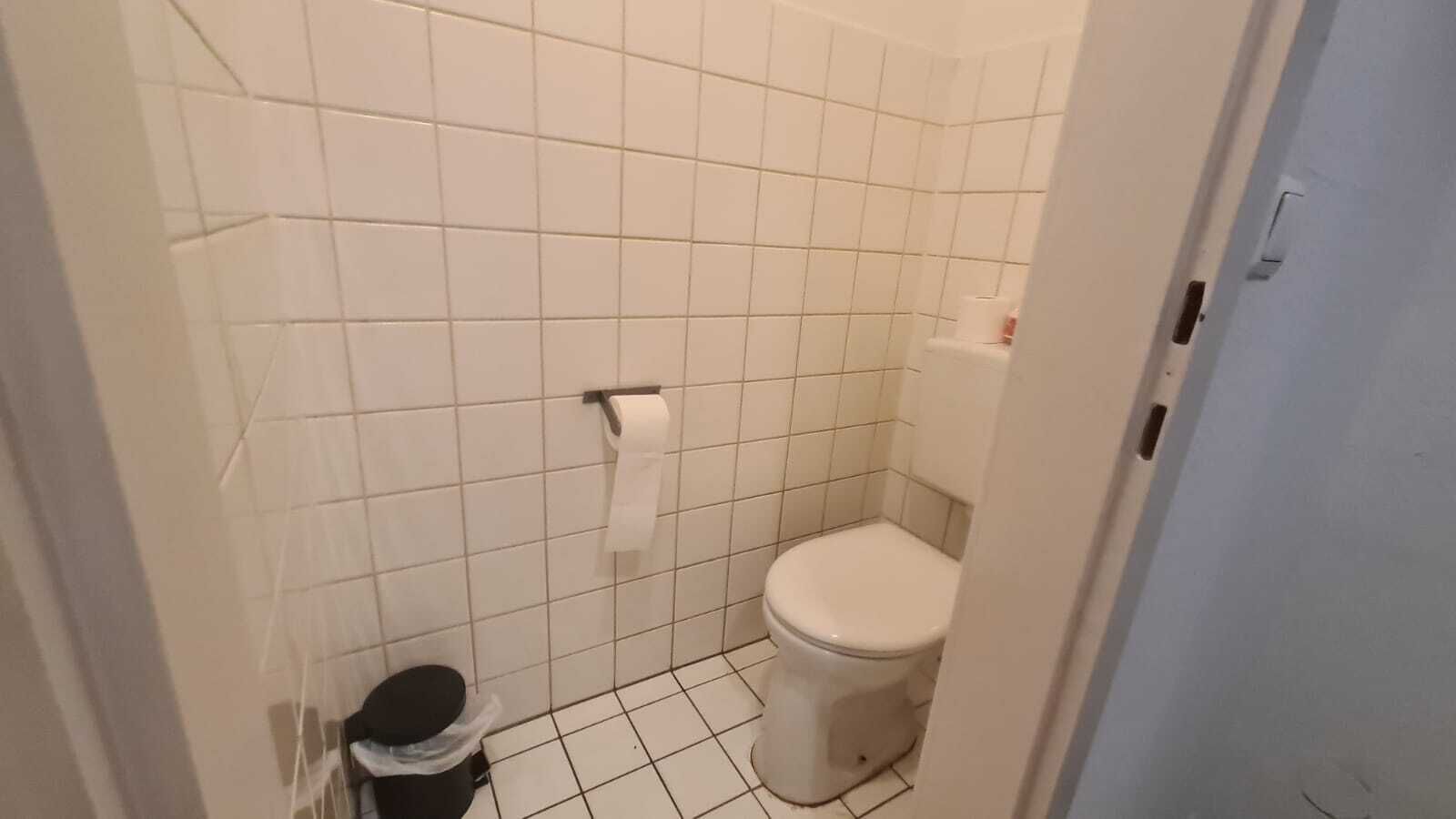 A small, tiled bathroom with a toilet, a trash bin, and a toilet paper holder.