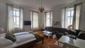 Ein Hotelzimmer mit zwei Betten, einem Schlafsofa, einem Couchtisch, einem Fernseher und einem Fenster mit Vorhängen.
