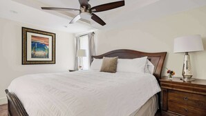Une chambre d’hôtel avec un grand lit, un ventilateur de plafond, une table de chevet avec une lampe, un tableau encadré au mur et une fenêtre avec des rideaux.