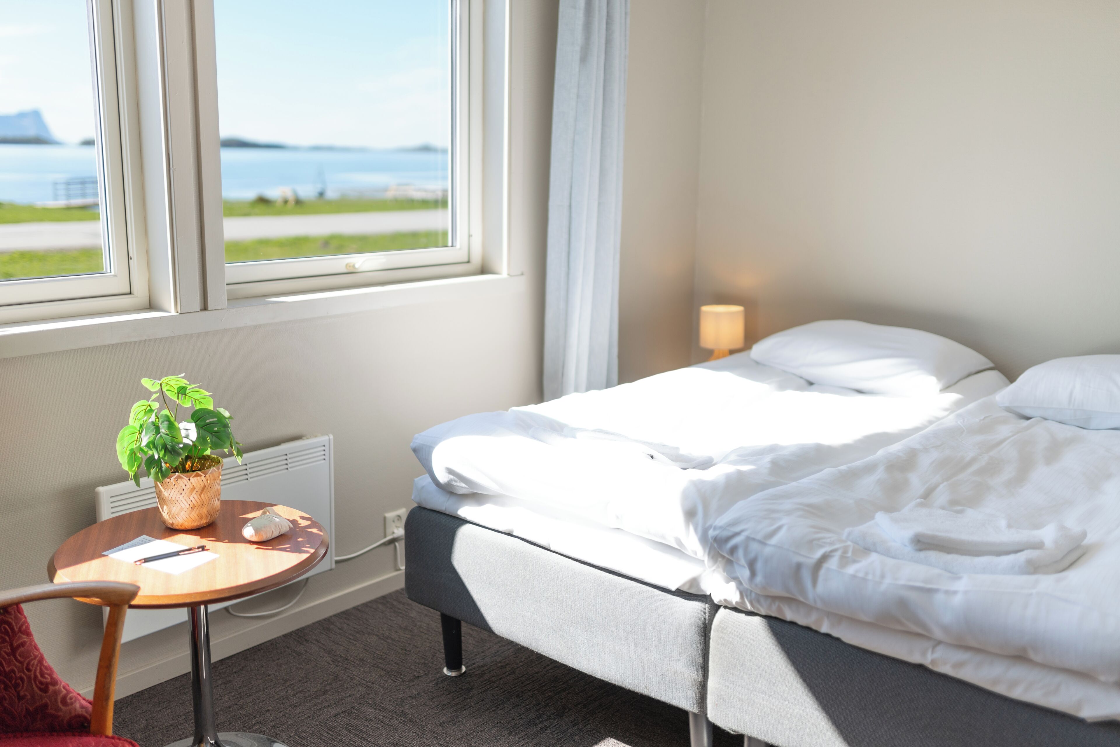 Ett hotellrum med en säng, ett litet bord med en växt, en lampa och utsikt över havet genom ett fönster.