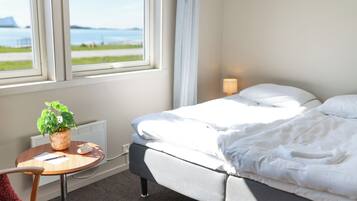 Ett hotellrum med en säng, ett litet bord med en växt, en lampa och utsikt över havet genom ett fönster.