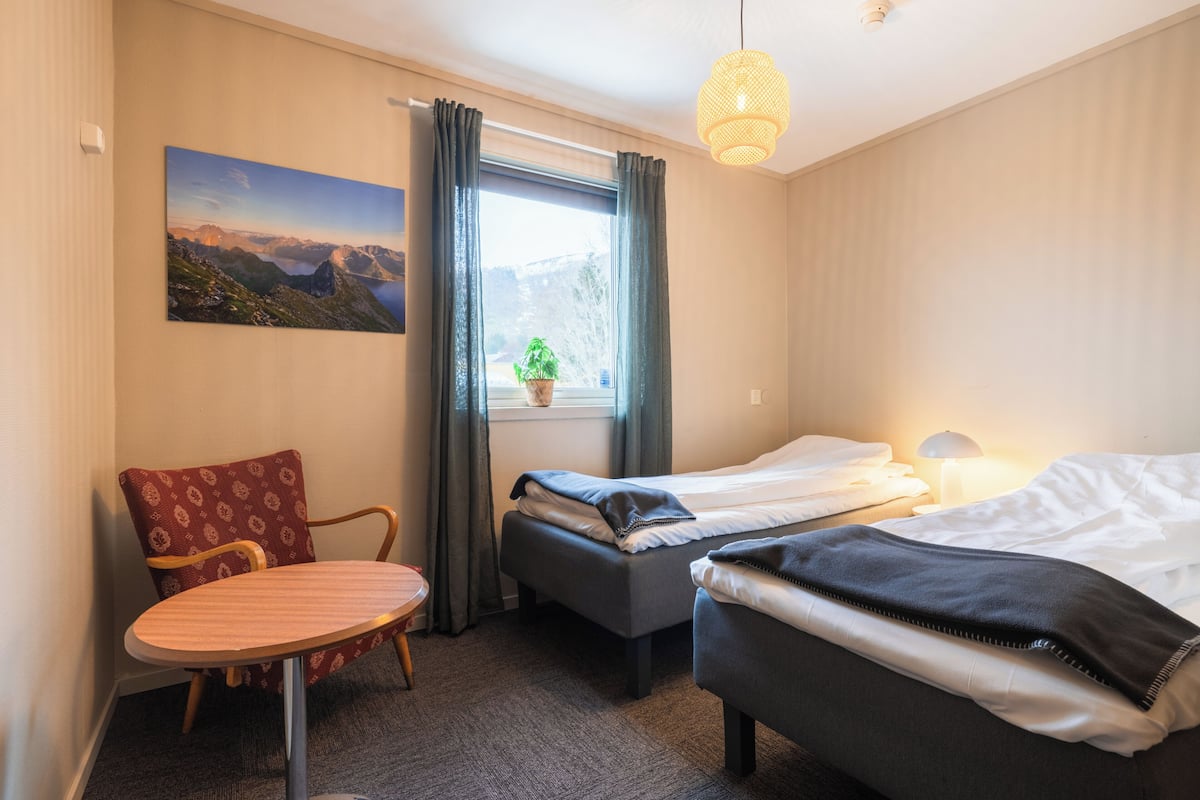 Et hotelværelse med seng, stol, lille bord, et vindue med gardiner, en plante på vindueskarmen og et billede på væggen.
