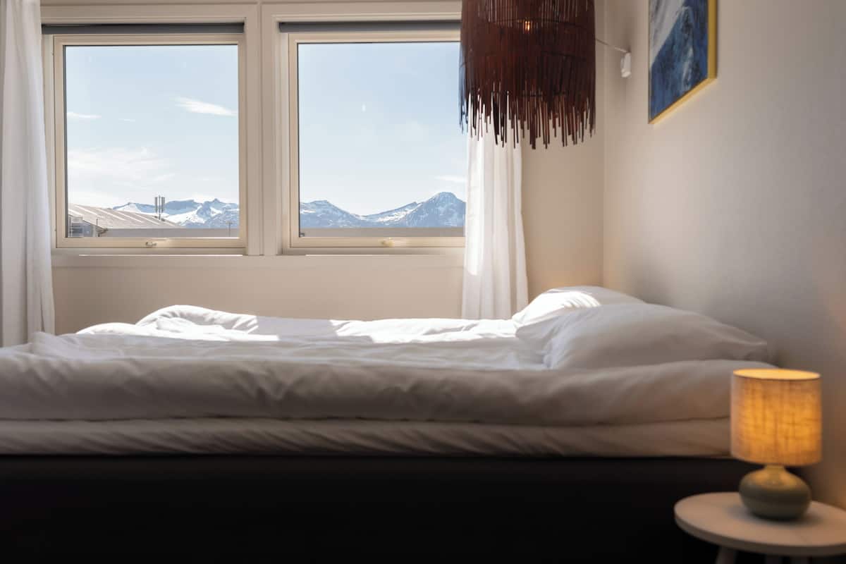 En seng med hvide sengetøj, en sengetårnslampe og et vindue med udsigt til bjerge.