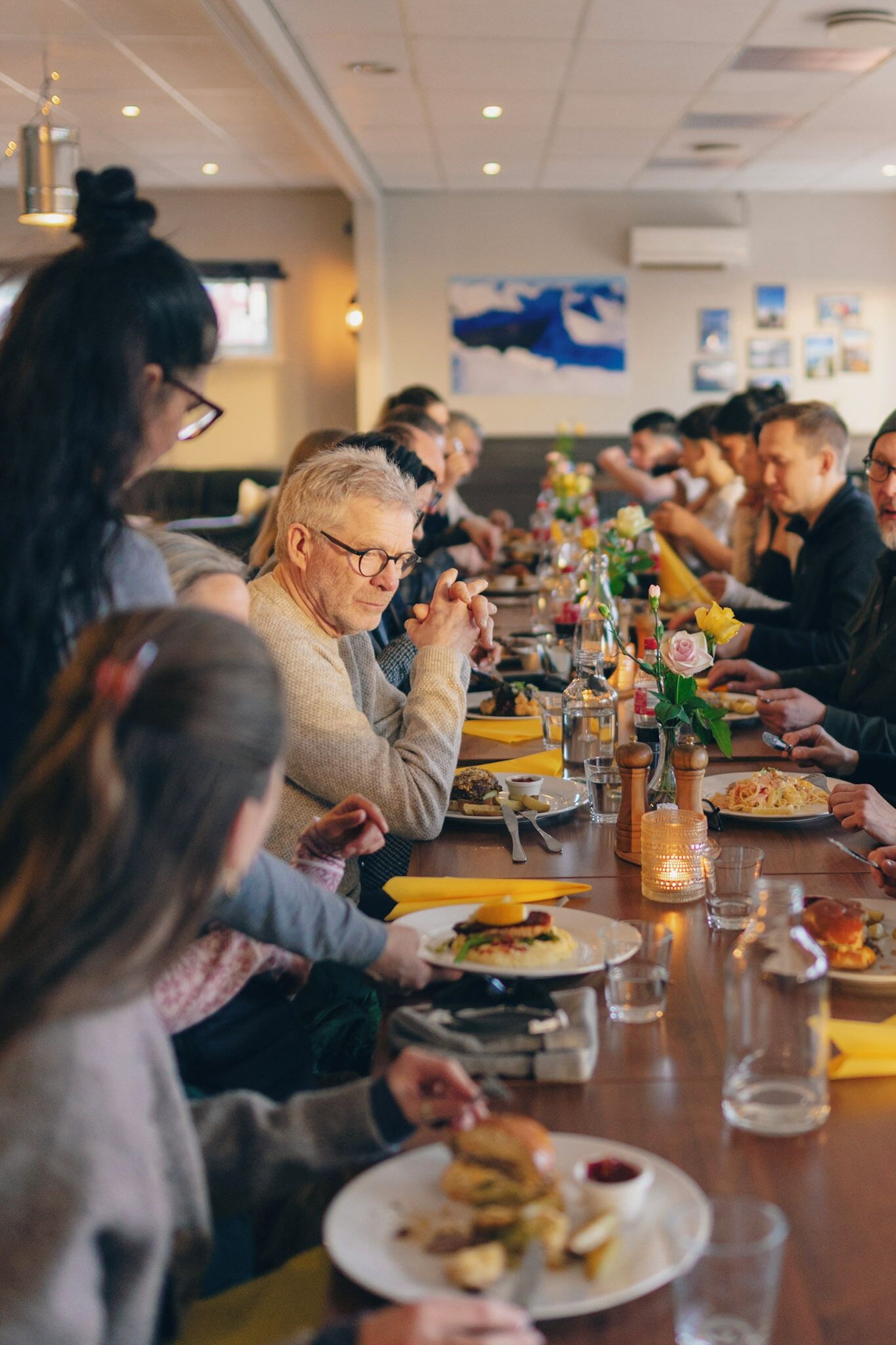 En grupp människor som äter middag på en restaurang, vid ett långt bord, med olika rätter och en dekorativ bordsdekoration i mitten.