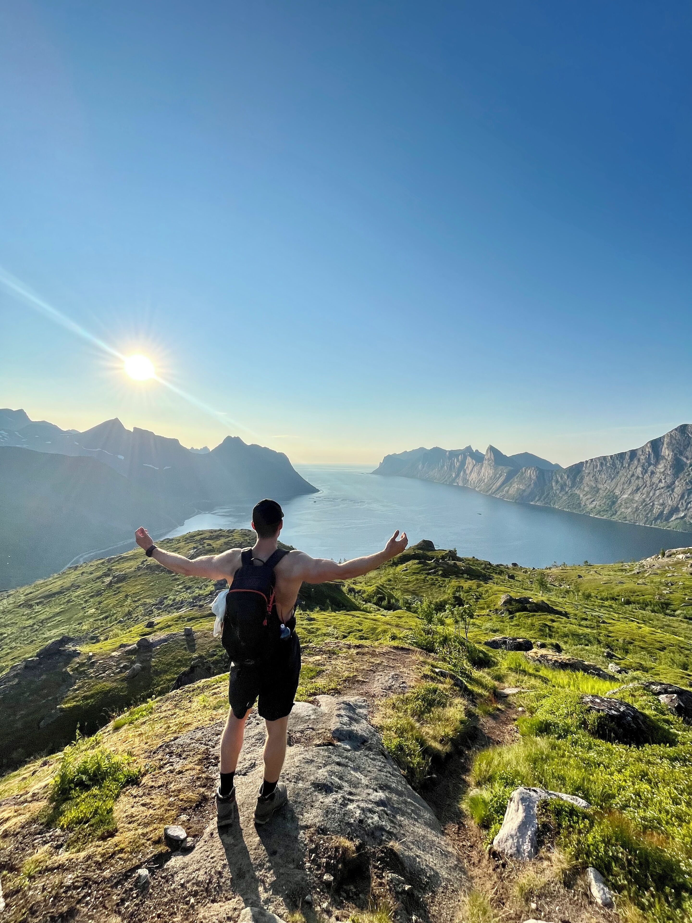 A person standing on a rocky outcrop with arms outstretched, overlooking a mountainous landscape and a body of water.
