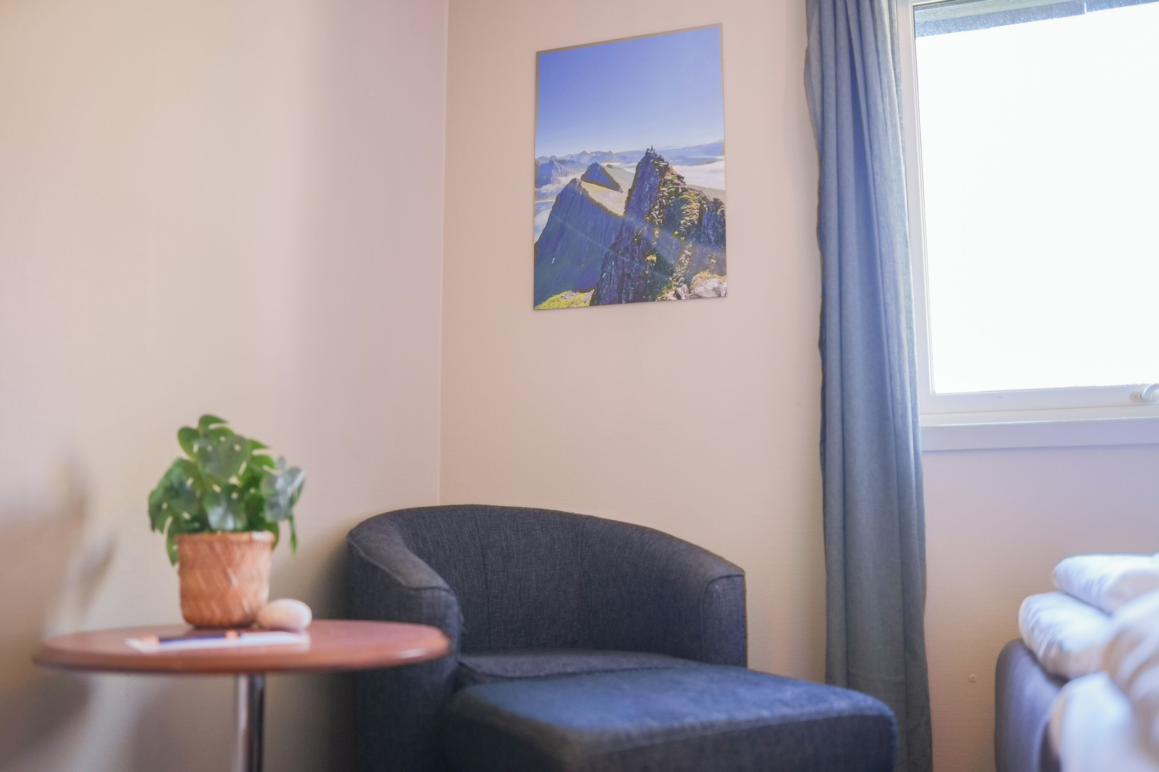 A room with a dark armchair, a small table with a potted plant, a framed picture on the wall, and a window with curtains.