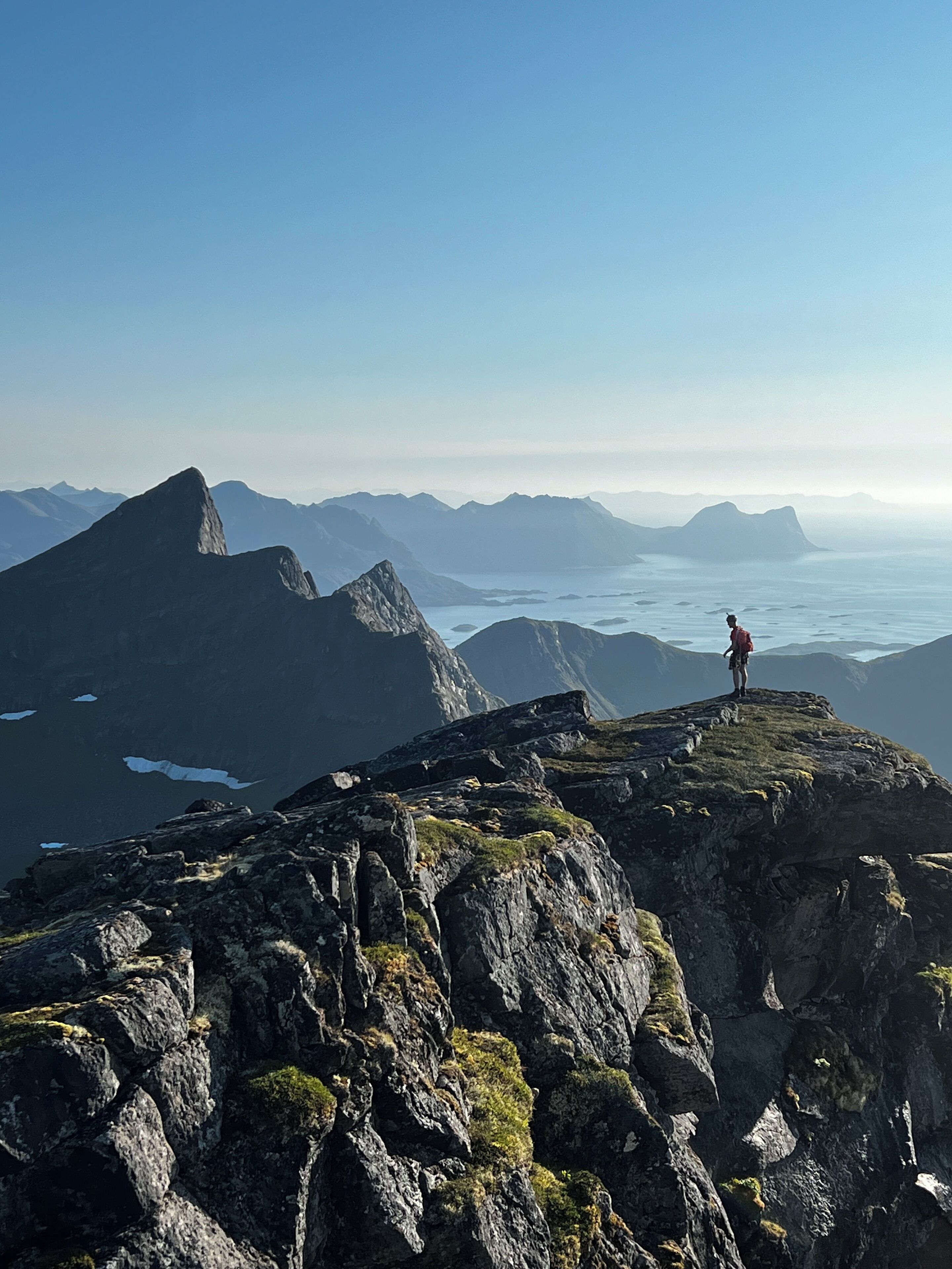 A person standing on a rocky mountain peak with a view of distant mountains and a body of water.