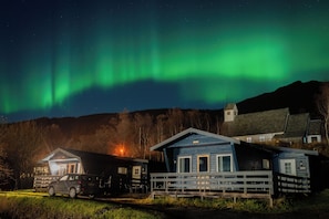 Una cabaña con un coche estacionado adelante, una valla de madera y vistas de las auroras boreales.
