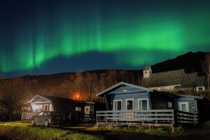 Una cabaña con un coche estacionado adelante, una valla de madera y vistas de las auroras boreales.