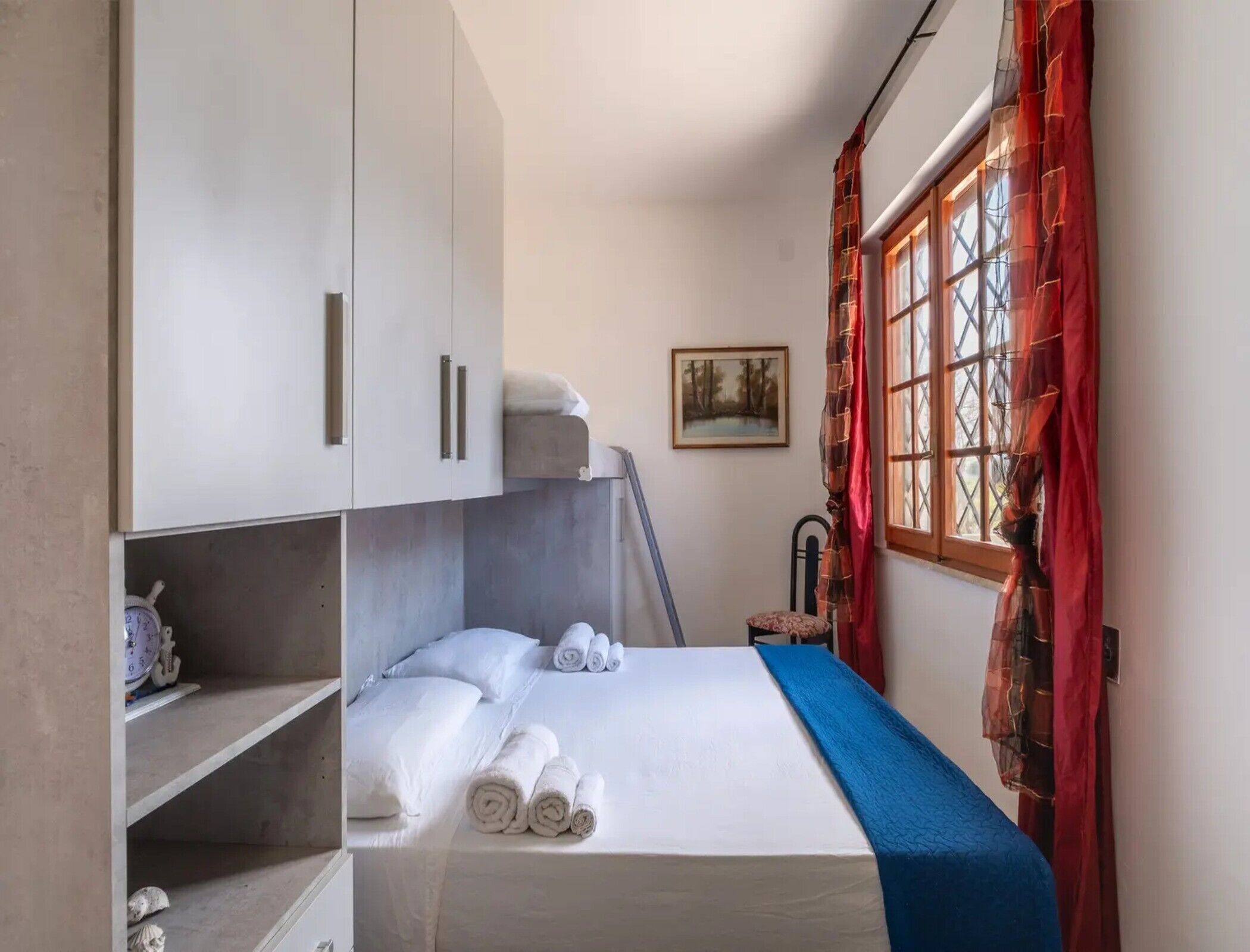 Ein Einzelbett mit blauer Bettdecke, einem weißen Kopfteil und einem Fenster mit roten Vorhängen.
