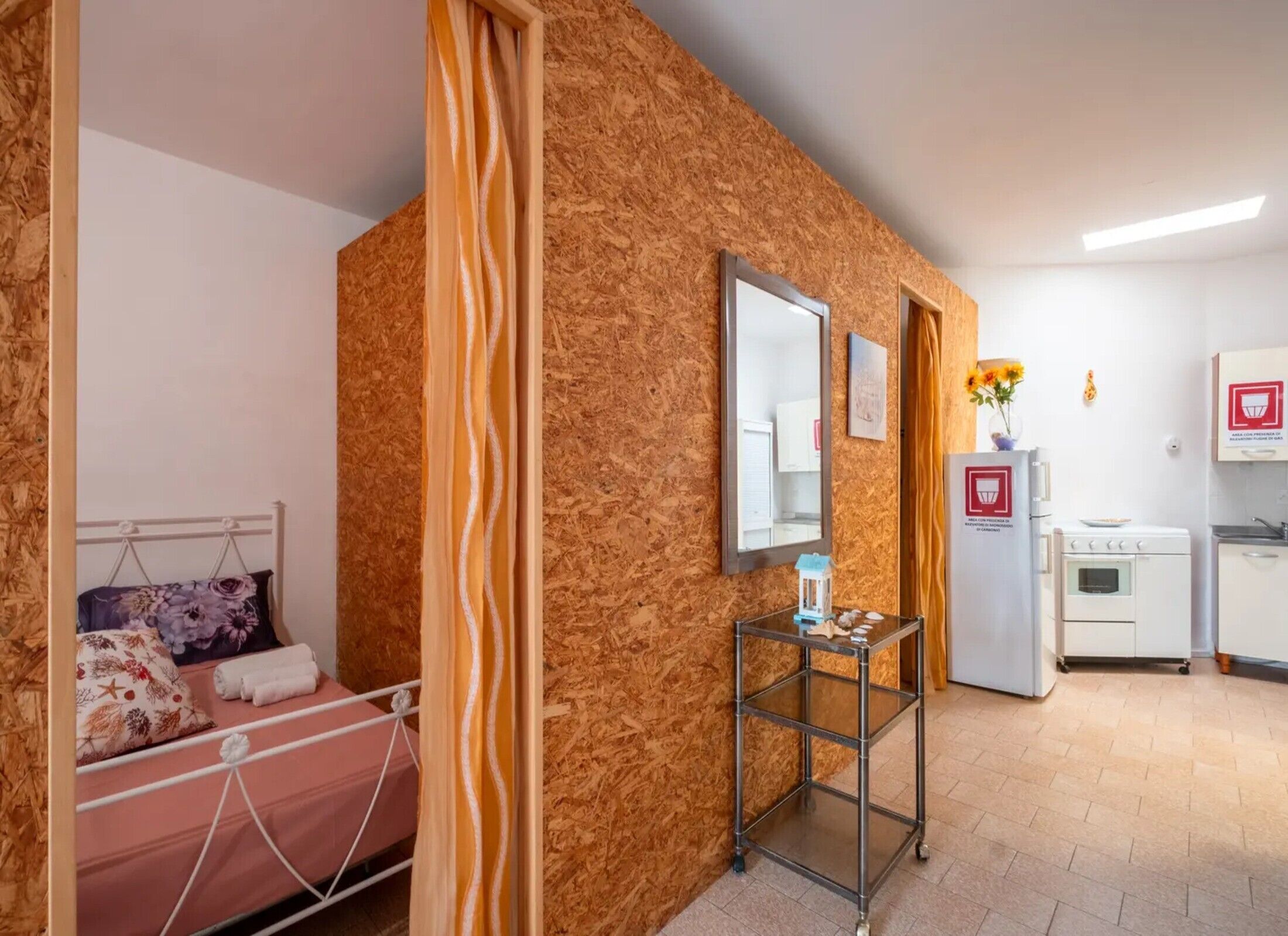 Una stanza con un letto, uno specchio, un tavolino con un vaso e angolo cottura.