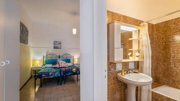 Een slaapkamer met een bed, nachtkastjes en een badkamer met een wastafel, spiegel en douche.