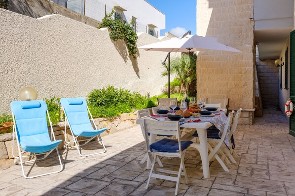 An outdoor patio with a table set for dining, two blue lounge chairs, and a white umbrella.