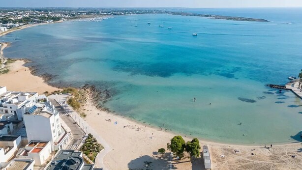 Luchtbeeld van een kustgebied met een zandstrand, helderblauw water en een verre kustlijn met gebouwen.